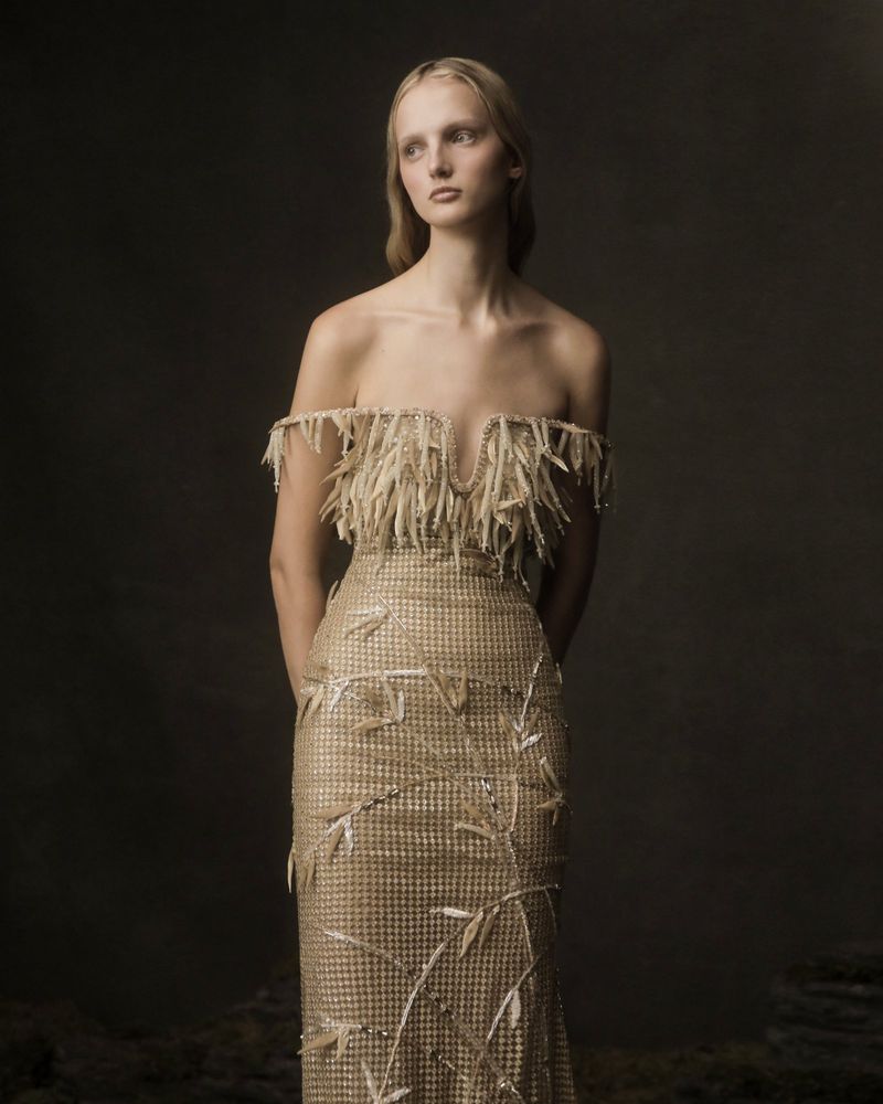 A fair-skinned woman with long blonde hair models a gold off-the-shoulder gown with tassels and floral accents against a dark studio backdrop.