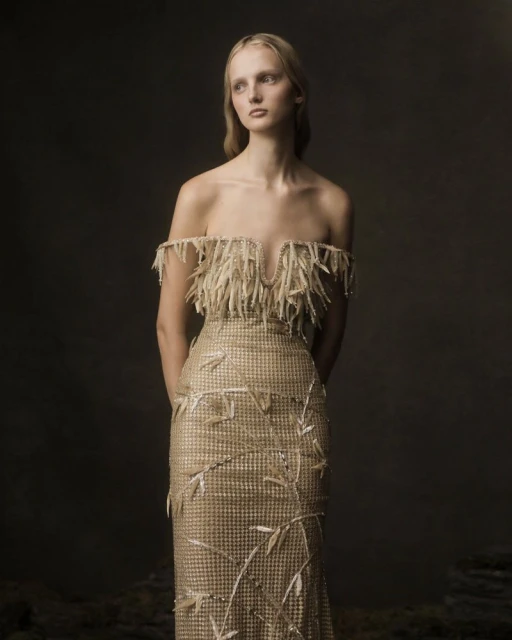 A fair-skinned woman with long blonde hair models a gold off-the-shoulder gown with tassels and floral accents against a dark studio backdrop.
