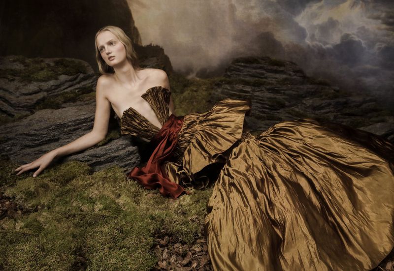 Woman in golden gown reclines on mossy ground with rocks and a cloudy sky backdrop in a fashion editorial, creating a surreal scene.