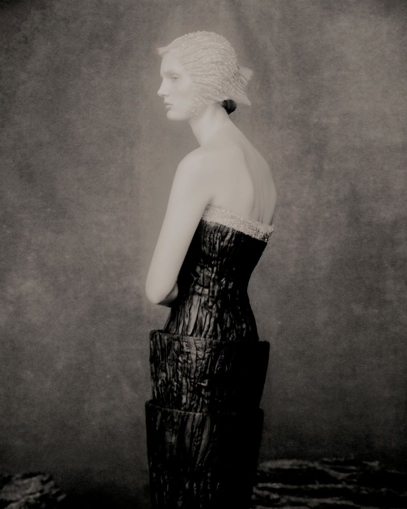 Monochrome studio shot of an adult female fashion model in profile, wearing a structured dress and hat, against a mottled backdrop.