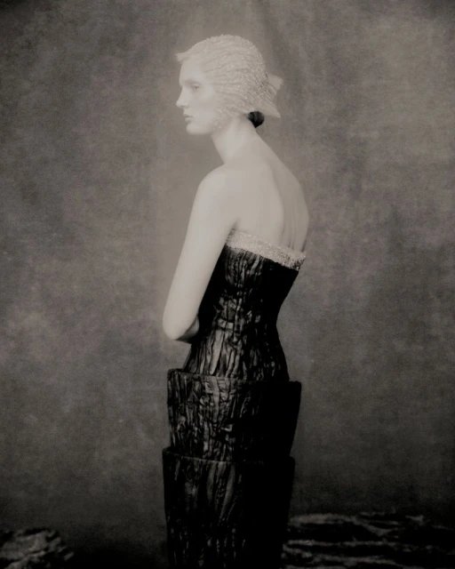 Monochrome studio shot of an adult female fashion model in profile, wearing a structured dress and hat, against a mottled backdrop.