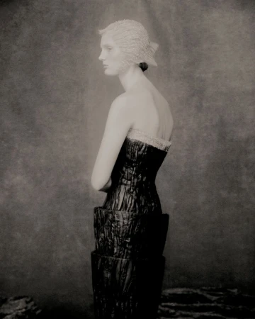 Monochrome studio shot of an adult female fashion model in profile, wearing a structured dress and hat, against a mottled backdrop.