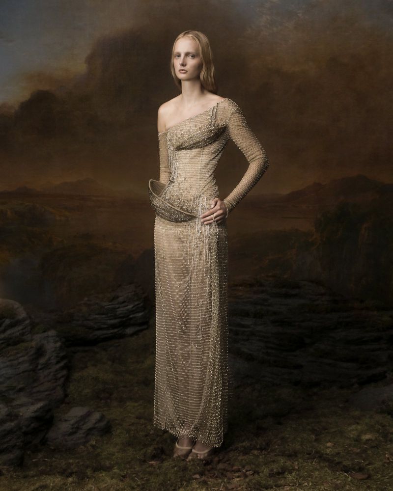Full-length shot of a blonde adult female wearing a gold beaded gown with landscape background, fashion editorial.