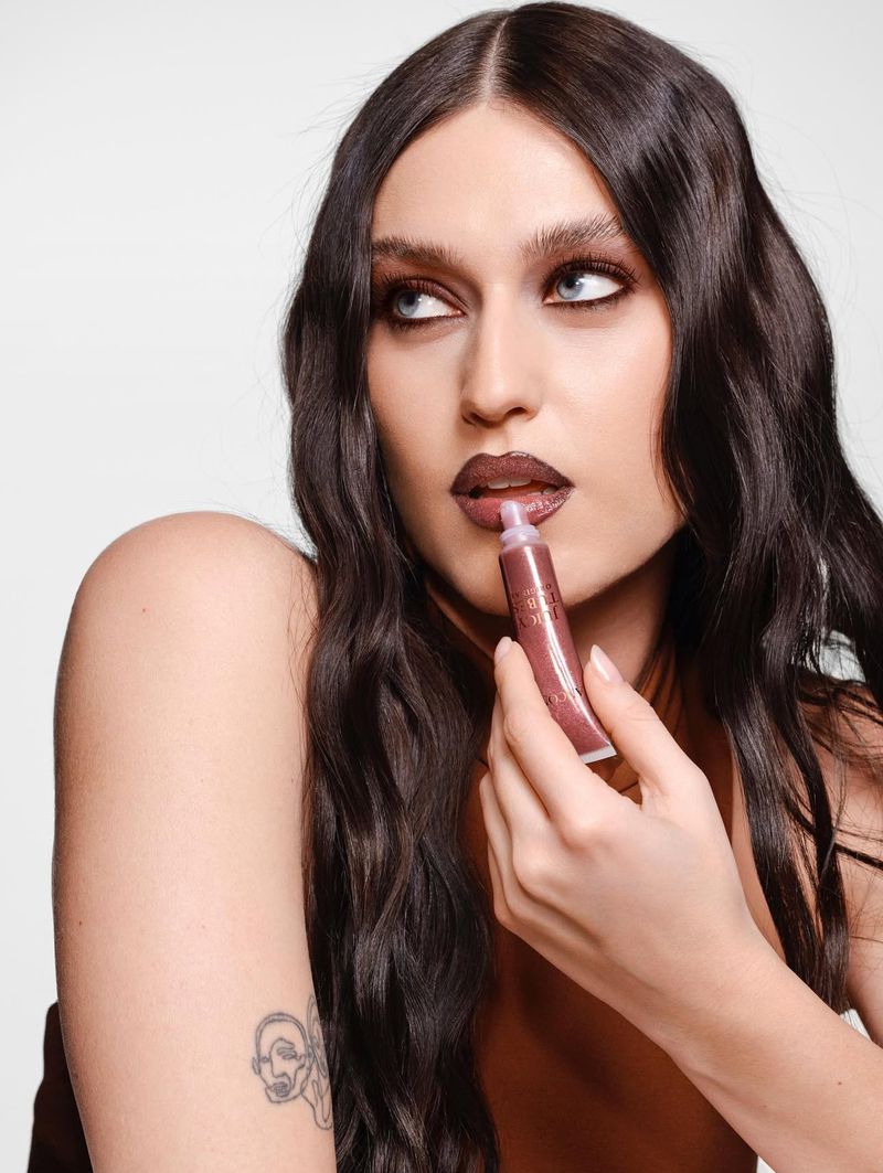 Close-up of an adult female applying lip gloss while looking away against a white backdrop in a beauty advertisement.