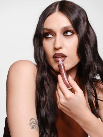 Close-up of an adult female applying lip gloss while looking away against a white backdrop in a beauty advertisement.