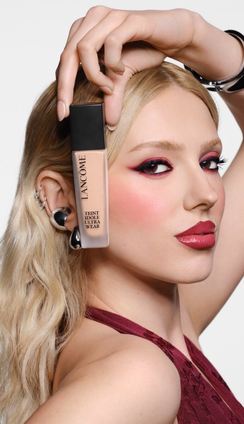 Woman holding Lancôme Teint Idole Ultra Wear foundation with makeup. She has blond hair and wears a sleeveless maroon top against a white background.