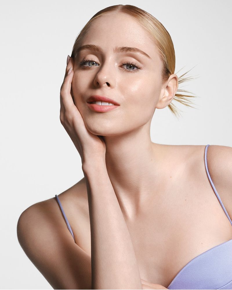 Close-up of a fair-skinned woman with blonde hair pulled back, wearing a purple tank top, posing against a clean white backdrop.