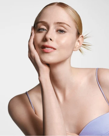 Close-up of a fair-skinned woman with blonde hair pulled back, wearing a purple tank top, posing against a clean white backdrop.