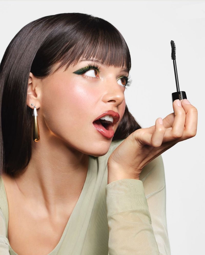 A young woman with dark hair and green eyeliner holds up mascara brush with open mouth, posing against white background. Beauty shot.
