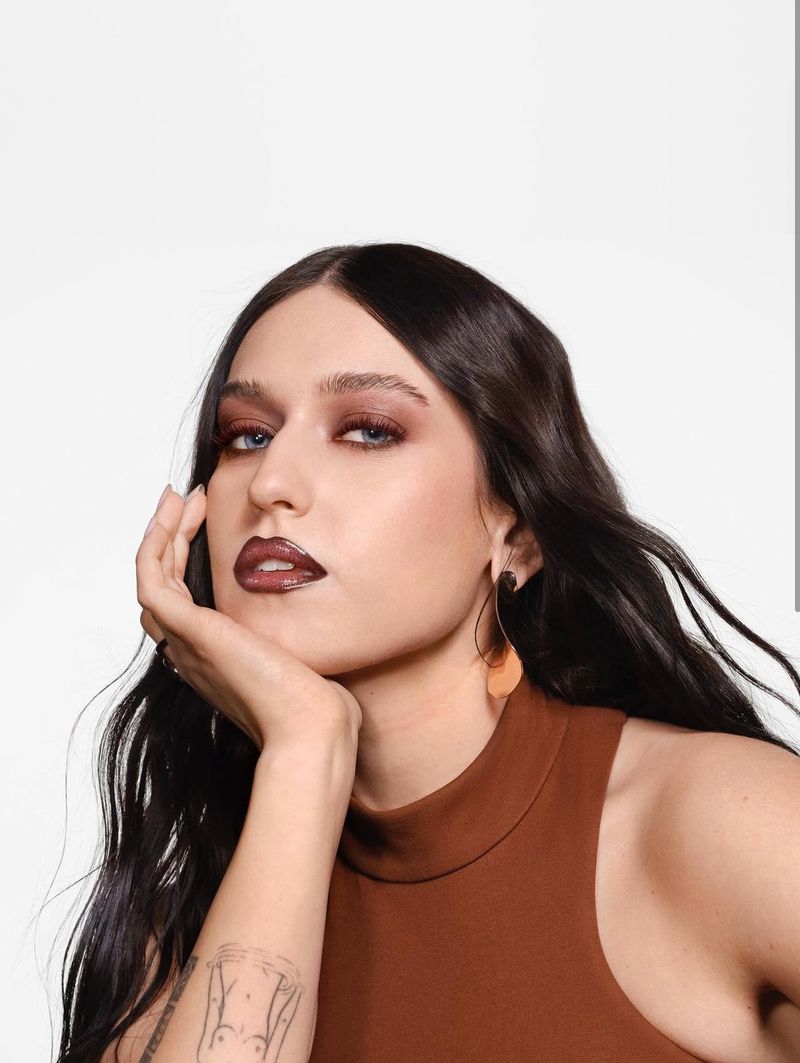 Close-up studio shot of a female adult with long wavy hair, wearing earrings, brown sleeveless top, with natural makeup and a tattoo on her arm.