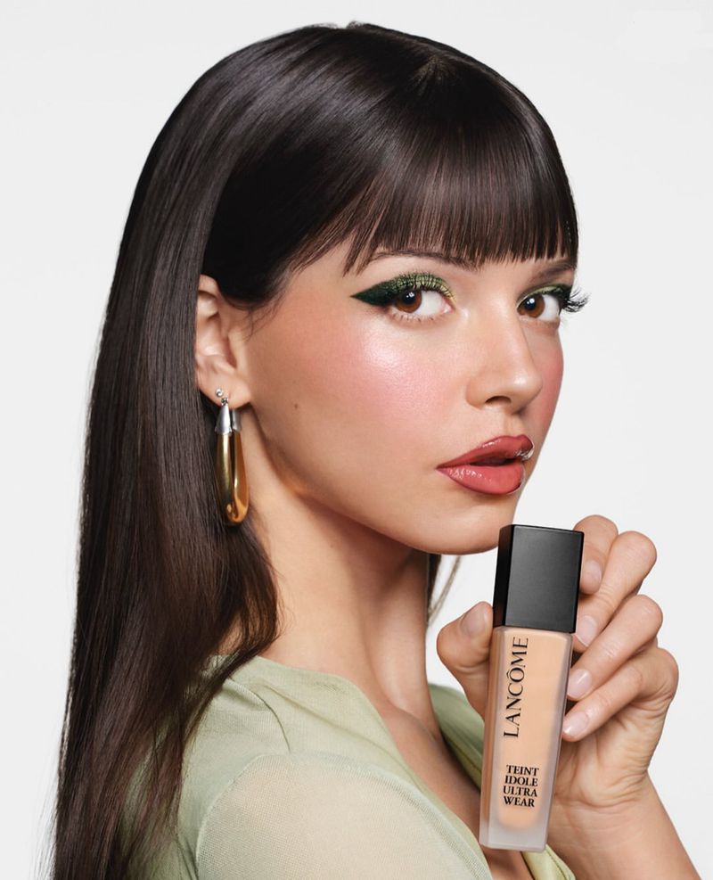 Brunette woman poses with Lancôme Teint Idole Ultra Wear foundation against white backdrop. Green eyeshadow winged eyeliner makeup look.