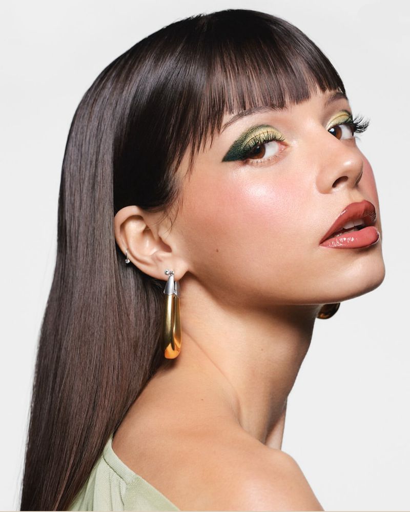 Close-up portrait of a young woman with brown hair, olive eyeshadow, and glossy lips, wearing hoop earrings against a white backdrop.