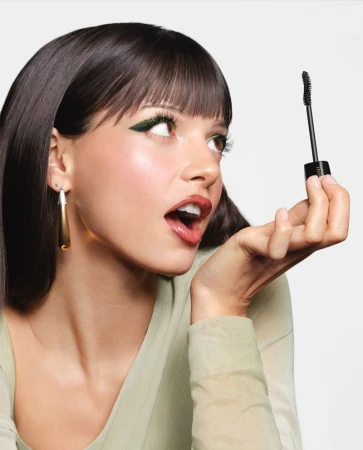 Woman with bangs and green eyeliner holding up a black mascara wand against a bright white background.