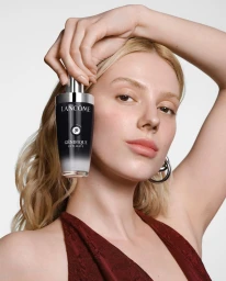 Blonde woman holding a black cosmetic bottle next to her face against a plain white background