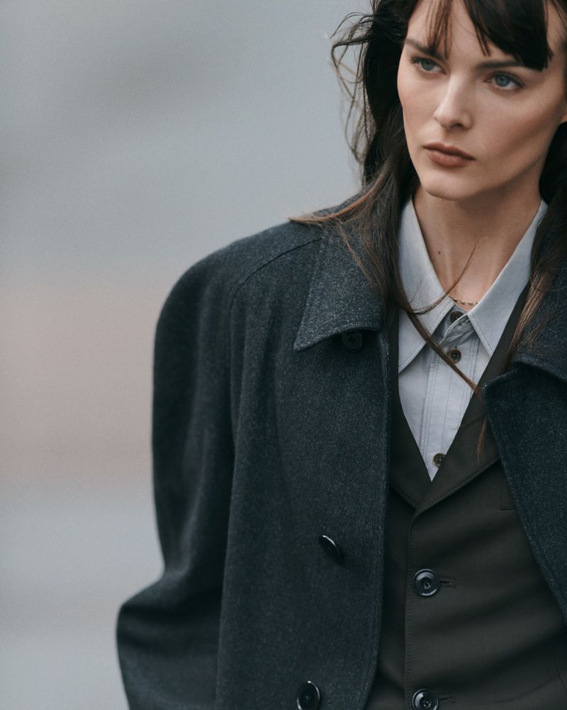 A stylish woman wearing a dark grey overcoat, vest and collared shirt gazes off camera in a medium shot with a blurred background.