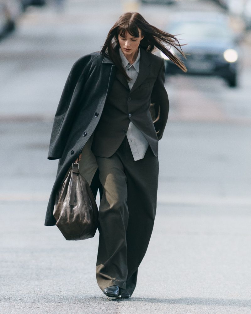 A woman in a suit and coat walks on a street with a car in the background, carrying a soft leather bag. The style is editorial.