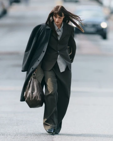 A woman in a suit and coat walks on a street with a car in the background, carrying a soft leather bag. The style is editorial.