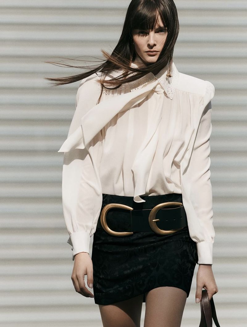 Woman in a white blouse and black skirt with a large belt, posing against a white wall, fashion editorial, studio shot.
