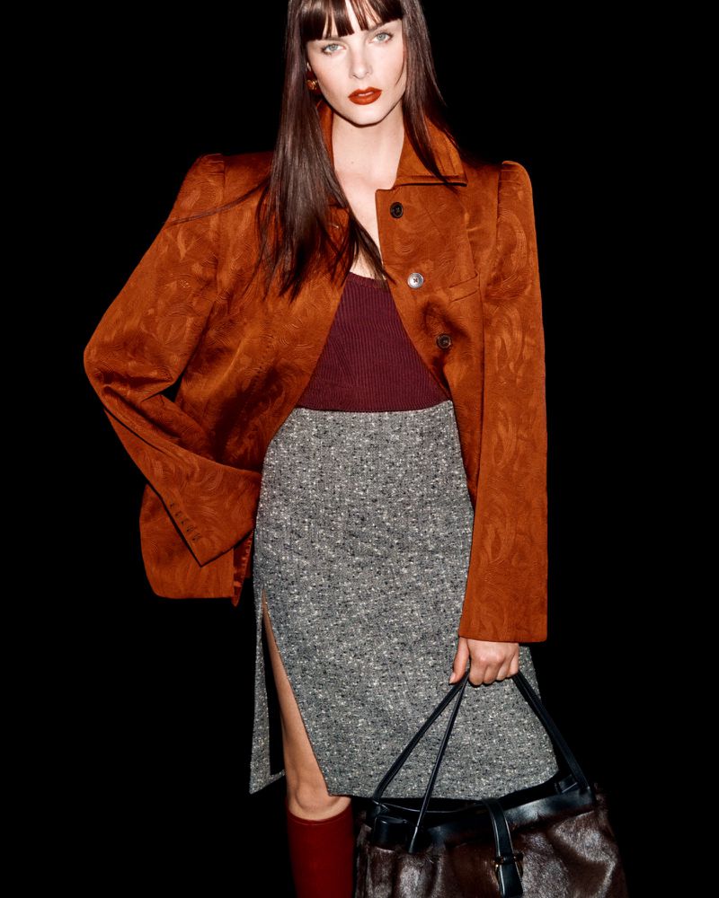 Fashionable adult woman poses in a brown jacket, grey skirt, and red boots against a black backdrop, carrying a handbag.