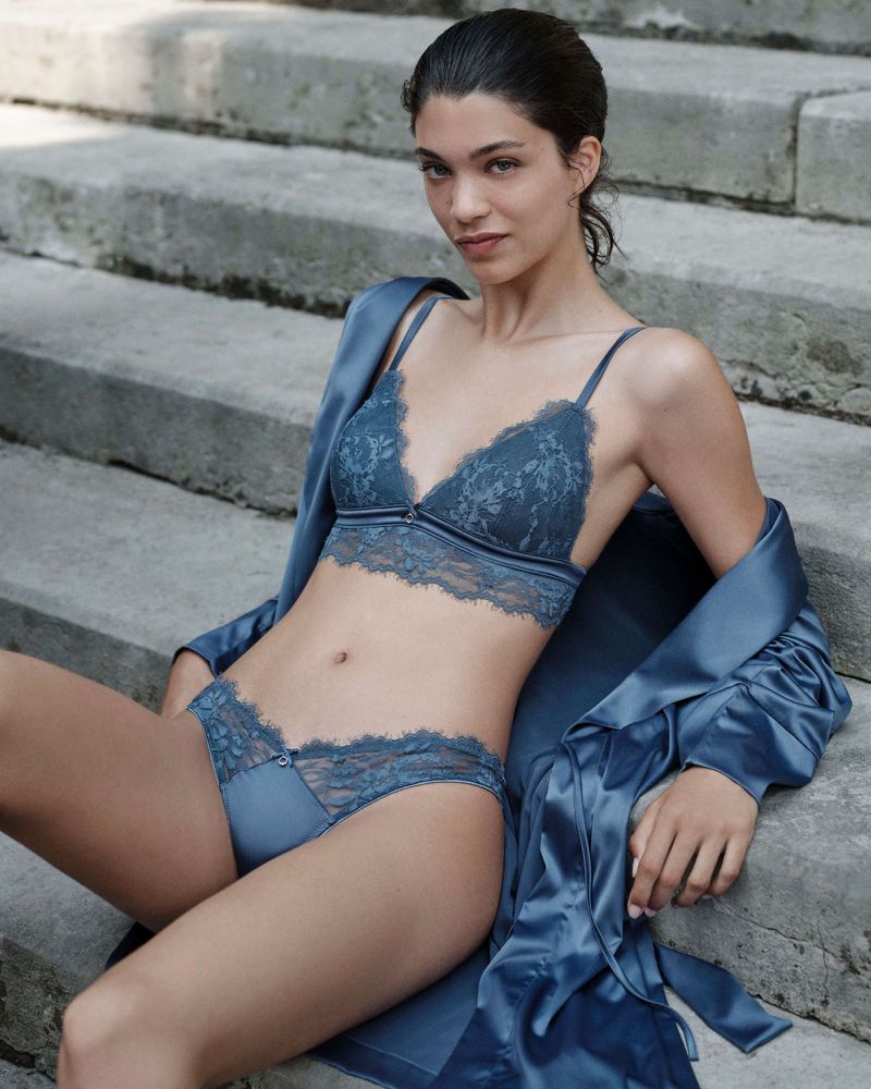 Brunette woman poses on stone steps wearing a blue lace bra and underwear set with a satin robe, capturing a sensual fashion moment.