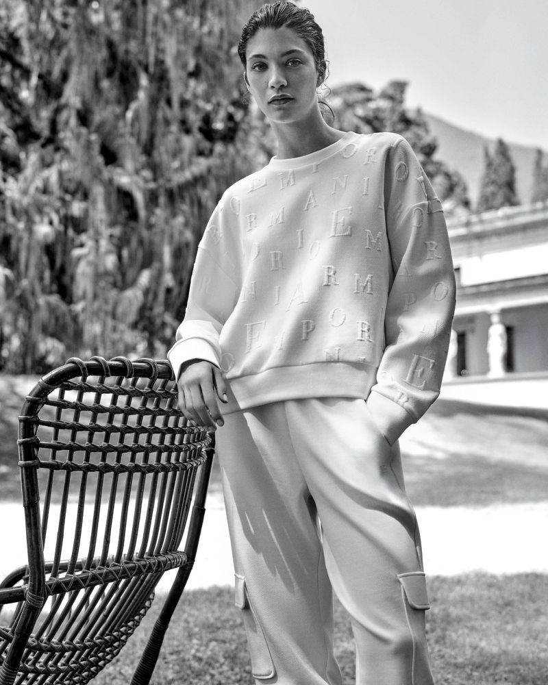 Monochrome portrait of a woman in an embossed Emporio Armani sweatshirt and sweatpants standing next to a wicker chair outdoors.