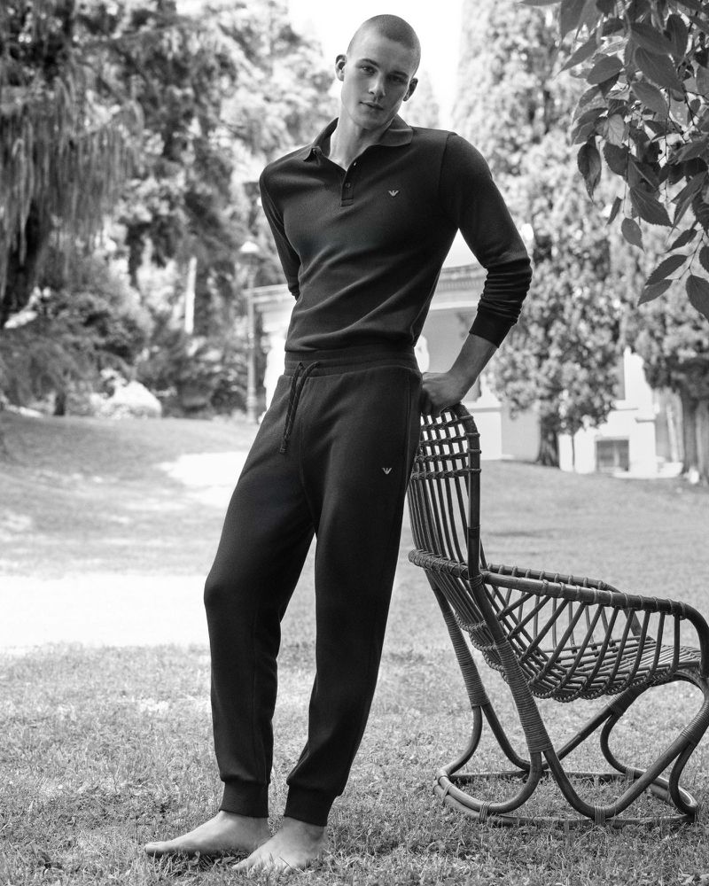 Monochrome full-length portrait of a barefoot man leaning against a wicker chair in a park setting.