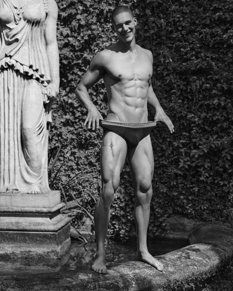 Black and white shot of a muscular adult male standing near a classical statue in an outdoor setting with lush green foliage.