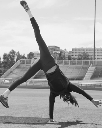 A person wearing workout clothes does a handstand on a green field with stadium seating and buildings in the background on a sunny day.