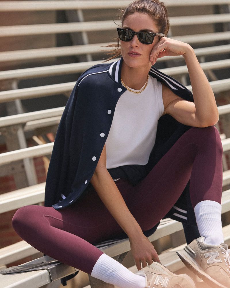 A woman in athletic wear sits on bleachers wearing sunglasses and a jacket, posing for a fitness or fashion shot on an overcast day.