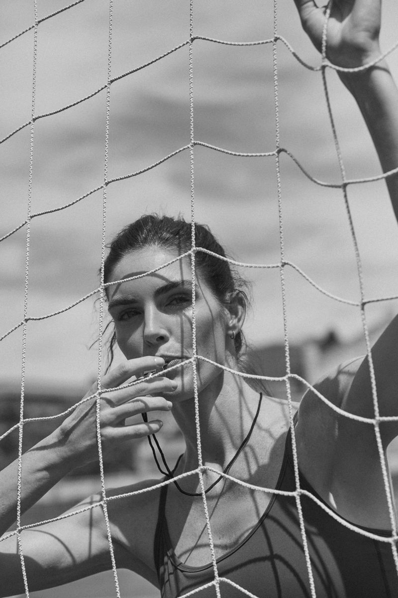 Monochrome portrait of a woman with a whistle, shot through a net. She poses with a neutral expression against a bright sky.