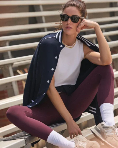 Model Hilary Rhoda wearing a navy bomber jacket and burgundy leggings seated on stadium bleachers for Tuckernuck
