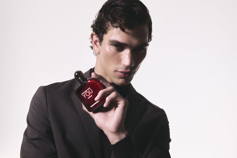 An adult male model wearing a black suit and turtleneck, holding a red cologne bottle against a bright white studio backdrop.