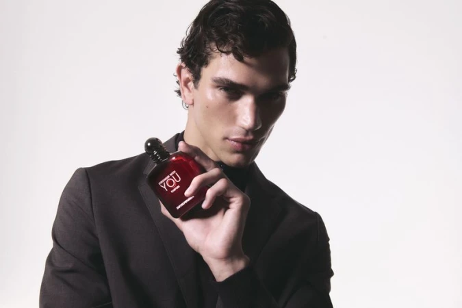 An adult male model wearing a black suit and turtleneck, holding a red cologne bottle against a bright white studio backdrop. 