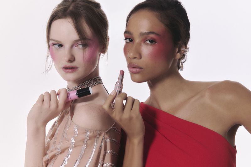Two women pose in a bright studio setting with makeup. One holds pink lipstick in front of a white backdrop, looking at camera.
