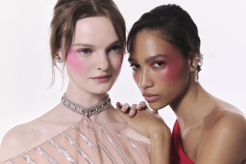 Two young women pose in a close-up studio shot, one with fair skin and the other with tan skin, both wearing minimal makeup.