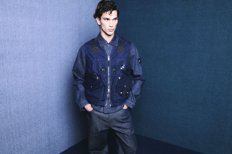 Male fashion model poses in a denim shirt and vest with hands in pockets against a blue textured background in a studio shot.
