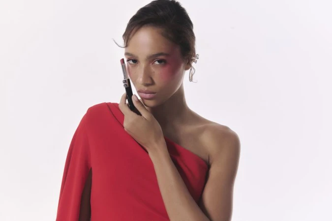 Model wearing a red one-shoulder dress holding a lipstick tube against a plain white background