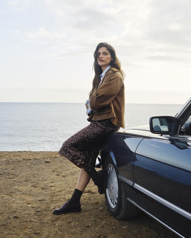 Brunette woman in a brown jacket and sequin skirt leaning against a black convertible with an ocean and sky background.