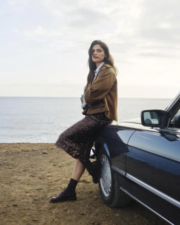 Brunette woman in a brown jacket and sequin skirt leaning against a black convertible with an ocean and sky background.