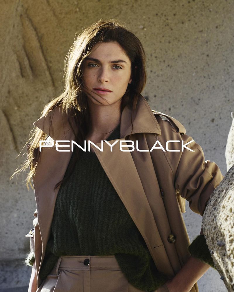 A woman with long brown hair poses in a studio wearing a brown trench coat and olive green sweater, with a rock formation background.