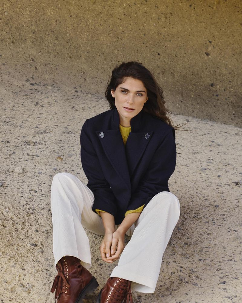 Woman in a navy pea coat and white pants sitting in front of a rock formation wearing brown lace-up boots in natural light.