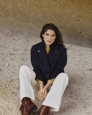 Woman in a navy pea coat and white pants sitting in front of a rock formation wearing brown lace-up boots in natural light.