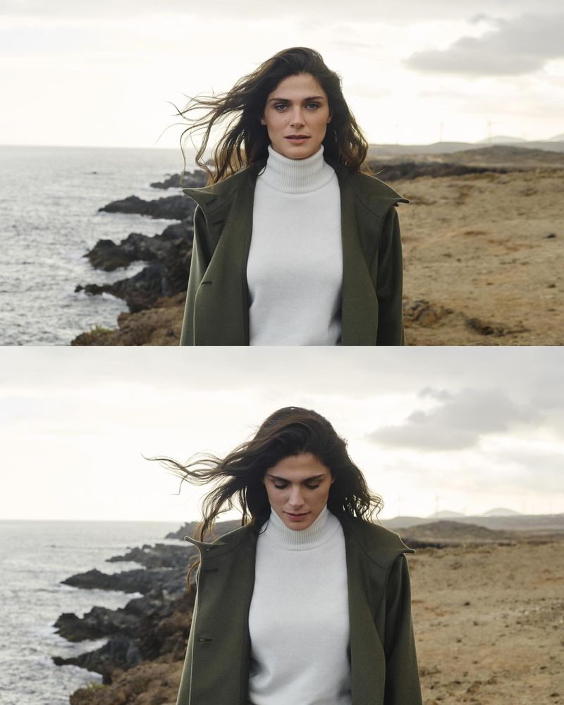 A woman with long, dark hair in a trench coat and turtleneck stands on a coastal cliff overlooking the ocean with cloudy skies in the background.