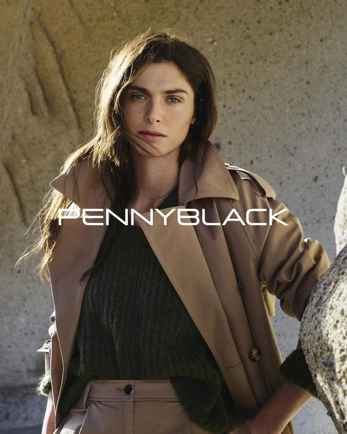 Model Elisa Sednaoui wearing a camel trench coat and green knit sweater against a rocky background for Pennyblack.