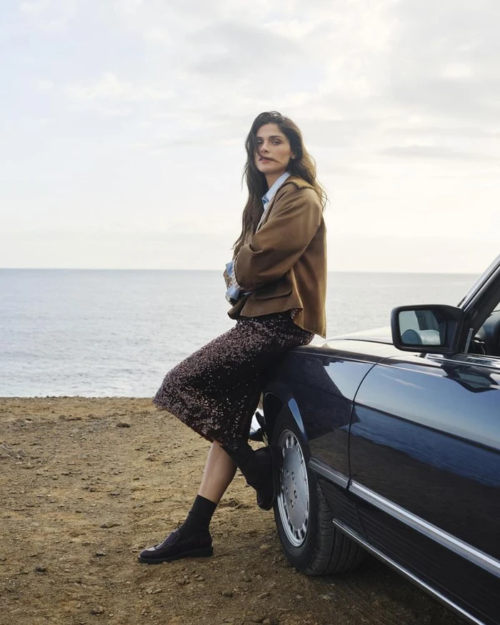 Model Elisa Sednaoui wearing a brown jacket and sequin skirt leaning against a dark car beside the ocean.
