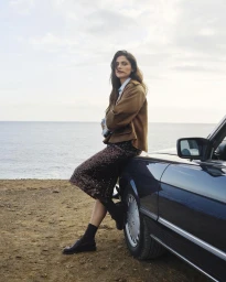 Model Elisa Sednaoui wearing a brown jacket and sequin skirt leaning against a dark car beside the ocean.
