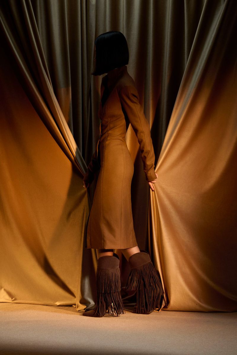 Brunette woman in tan dress and fringe boots standing in front of golden satin drapes. Fashion editorial shot. Indoor setting.