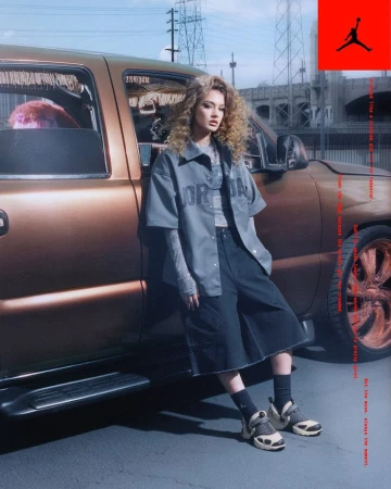 A female model poses by a gold pickup truck in an urban setting, wearing a Jordan shirt and dark denim shorts in an editorial fashion shot.