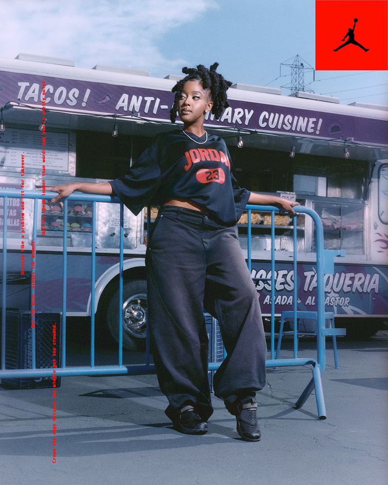 Young woman in streetwear poses in front of a taco truck. She wears a Jordan jersey, baggy jeans, and sneakers in an outdoor setting.