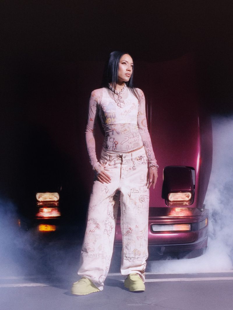 Woman in patterned top and pants poses in front of burgundy car with haze effect. Fashion shot, studio setting, diffused lighting.